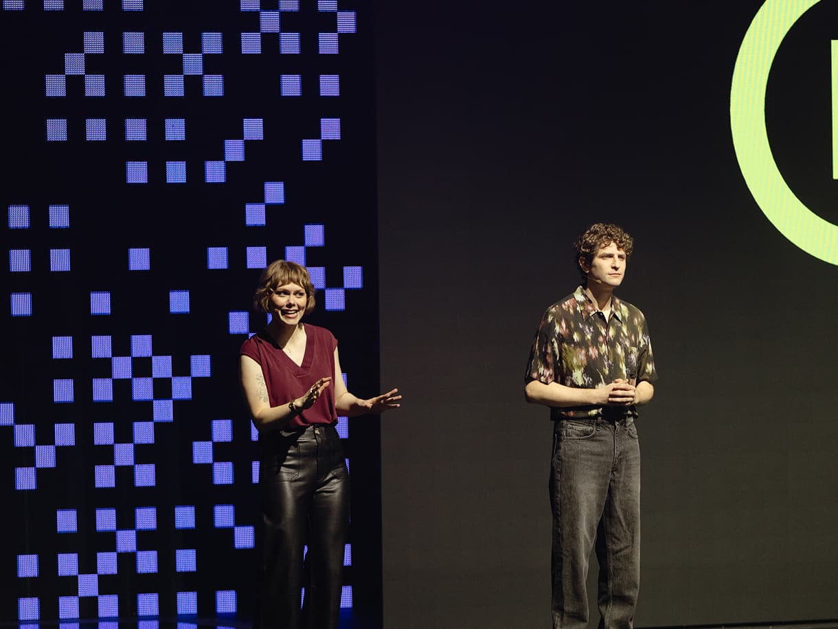 Two presenters on stage in front of a pixel-patterned video wall at dbt Summit 2026
